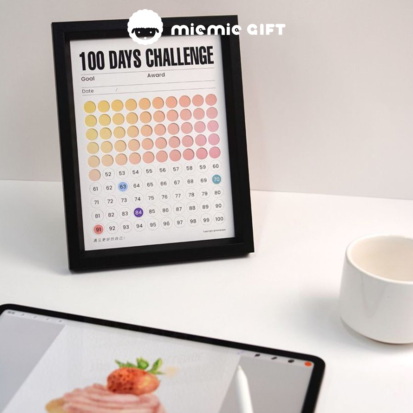 A black-framed 100 Days Challenge tracker with a gradient color design, placed on a desk alongside a tablet displaying a dessert image and a white coffee mug. The scene highlights the frame's versatility as both a motivational tool and a sophisticated decor piece for creative spaces.