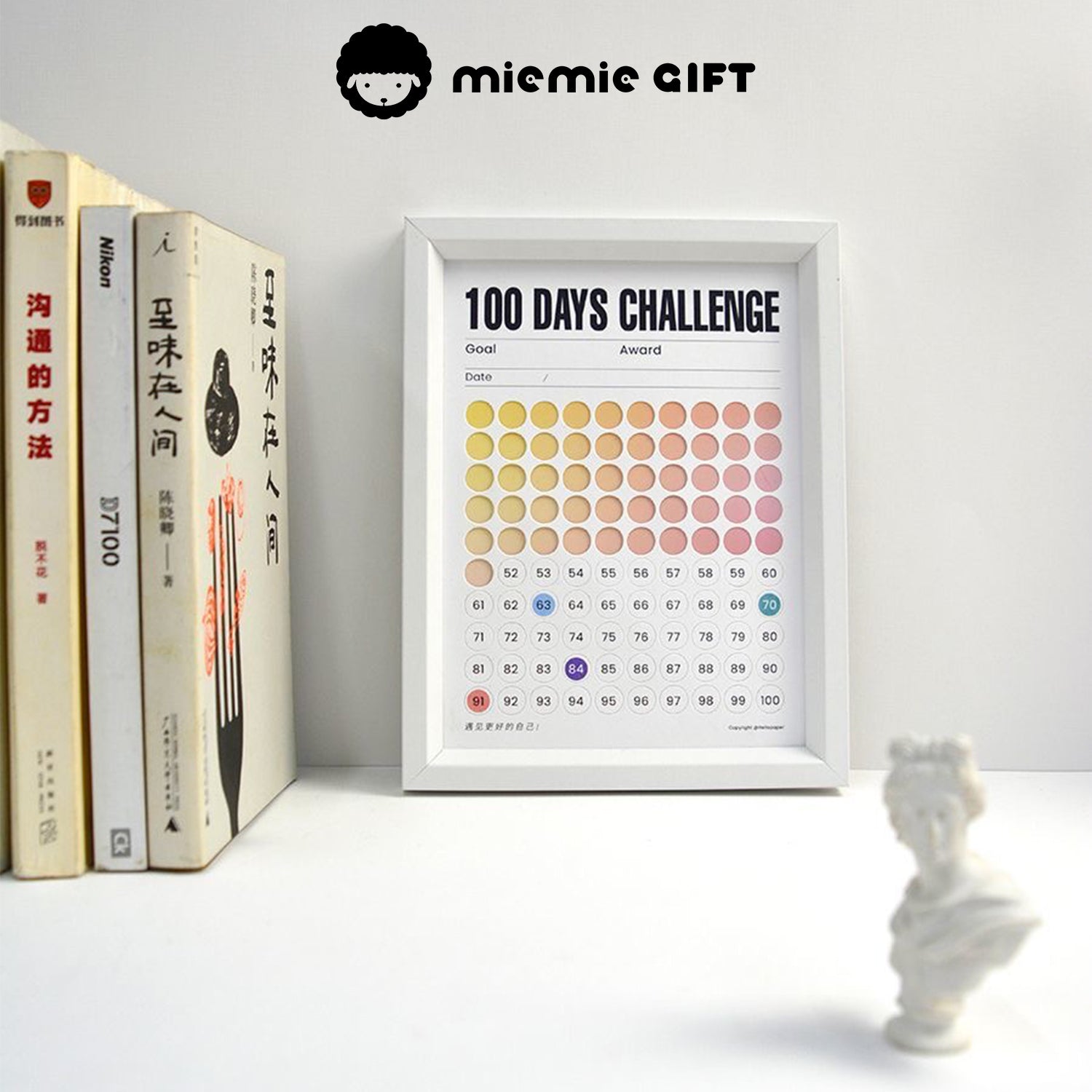 A white-framed 100 Days Challenge tracker with a gradient color design, placed on a desk alongside neatly arranged books and a miniature white sculpture. The frame combines functionality as a progress tracker with an elegant and inspiring workspace aesthetic.