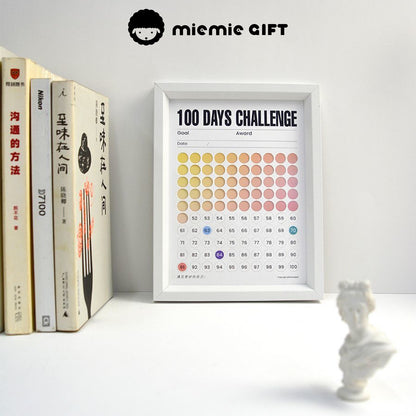 A white-framed 100 Days Challenge tracker with a gradient color design, placed on a desk alongside neatly arranged books and a miniature white sculpture. The frame combines functionality as a progress tracker with an elegant and inspiring workspace aesthetic.