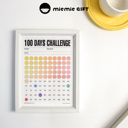 A white-framed 100 Days Challenge tracker with a gradient color design, displayed on a minimalist desk. The progress is visually marked, showcasing a motivational and practical approach to goal tracking. A yellow coaster with a white cup adds a touch of warmth to the scene.