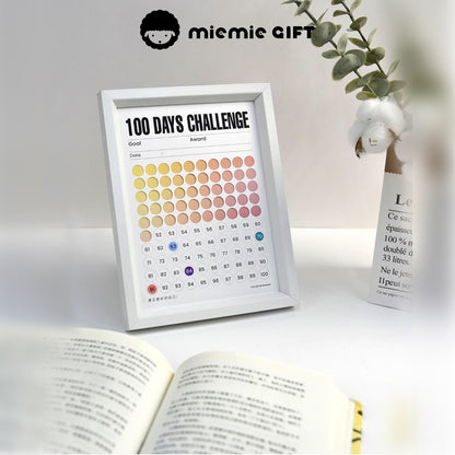 A white-framed 100 Days Challenge tracker with a gradient color design, displayed on a desk next to an open book and a minimalist vase with cotton branches. The frame highlights its functionality as a habit tracker and its aesthetic appeal for a peaceful and inspiring workspace.