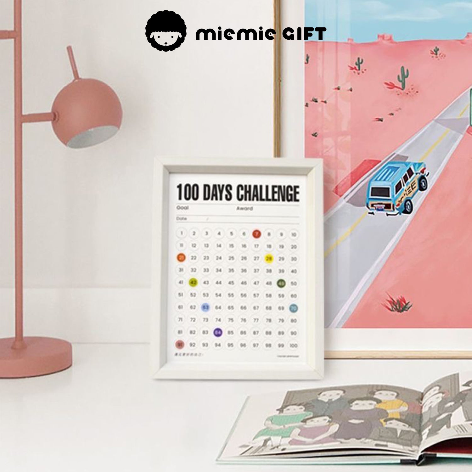 A white-framed 100 Days Challenge tracker placed on a clean desk, paired with a pink lamp and a colorful wall print depicting a road scene. The frame showcases its use as both a motivational tool and a stylish home decor piece, with a book opened nearby for a cozy and creative atmosphere.