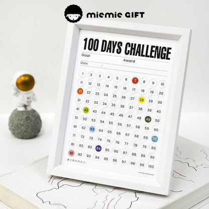 A white-framed 100 Days Challenge tracker placed on a desk, featuring numbered circles to mark daily progress. The frame is displayed alongside an astronaut figure, emphasizing motivation and creativity. Designed by MieMie Gift.