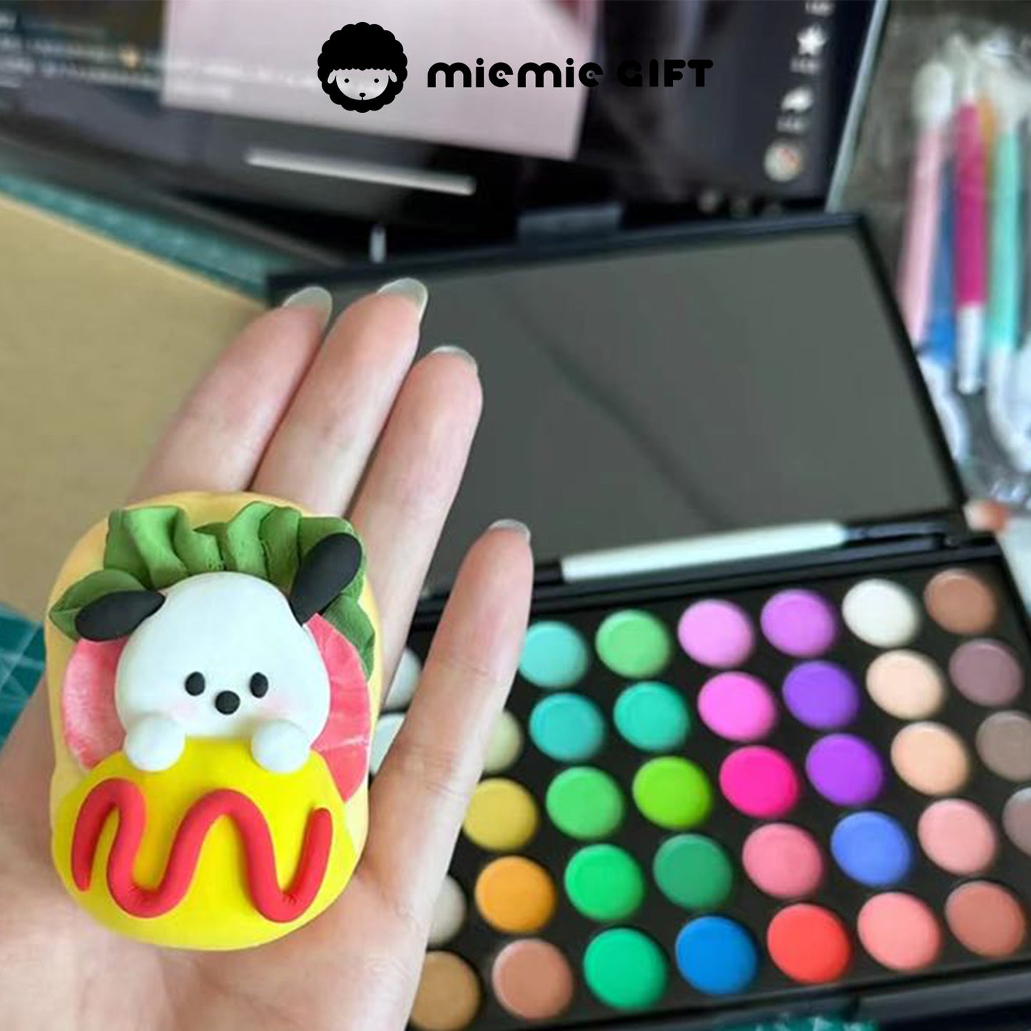 A hand holding an adorable handmade clay figurine of a puppy lying on a colorful dessert. The background features the MieMie Gift 40-Color Vivid Dream Palette, demonstrating the smooth and vibrant shading effect of the chalk powder on clay crafts.