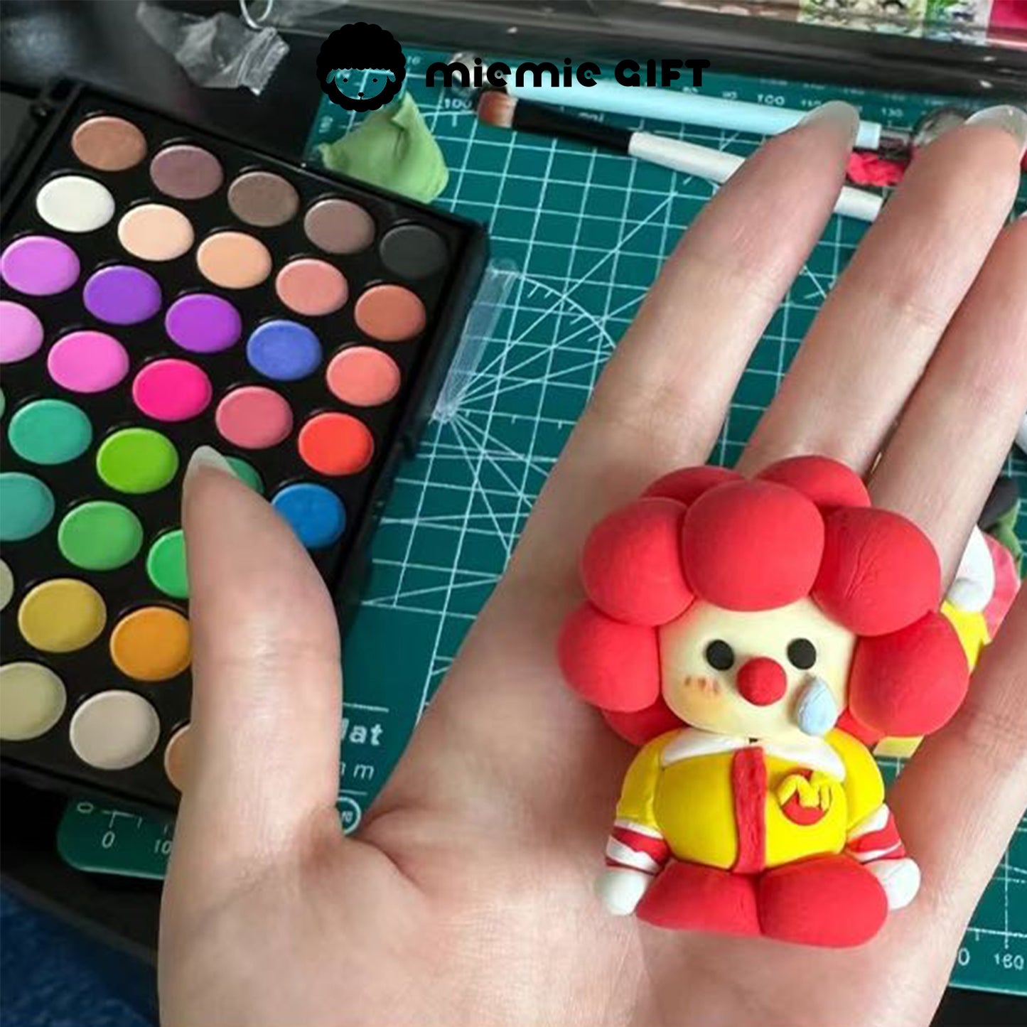 A hand holding a red curly-haired clown-style clay figurine. The background features the MieMie Gift 40-Color DIY Chalk Powder Palette, showcasing how the colors can bring clay crafts to life with rich and playful tones.