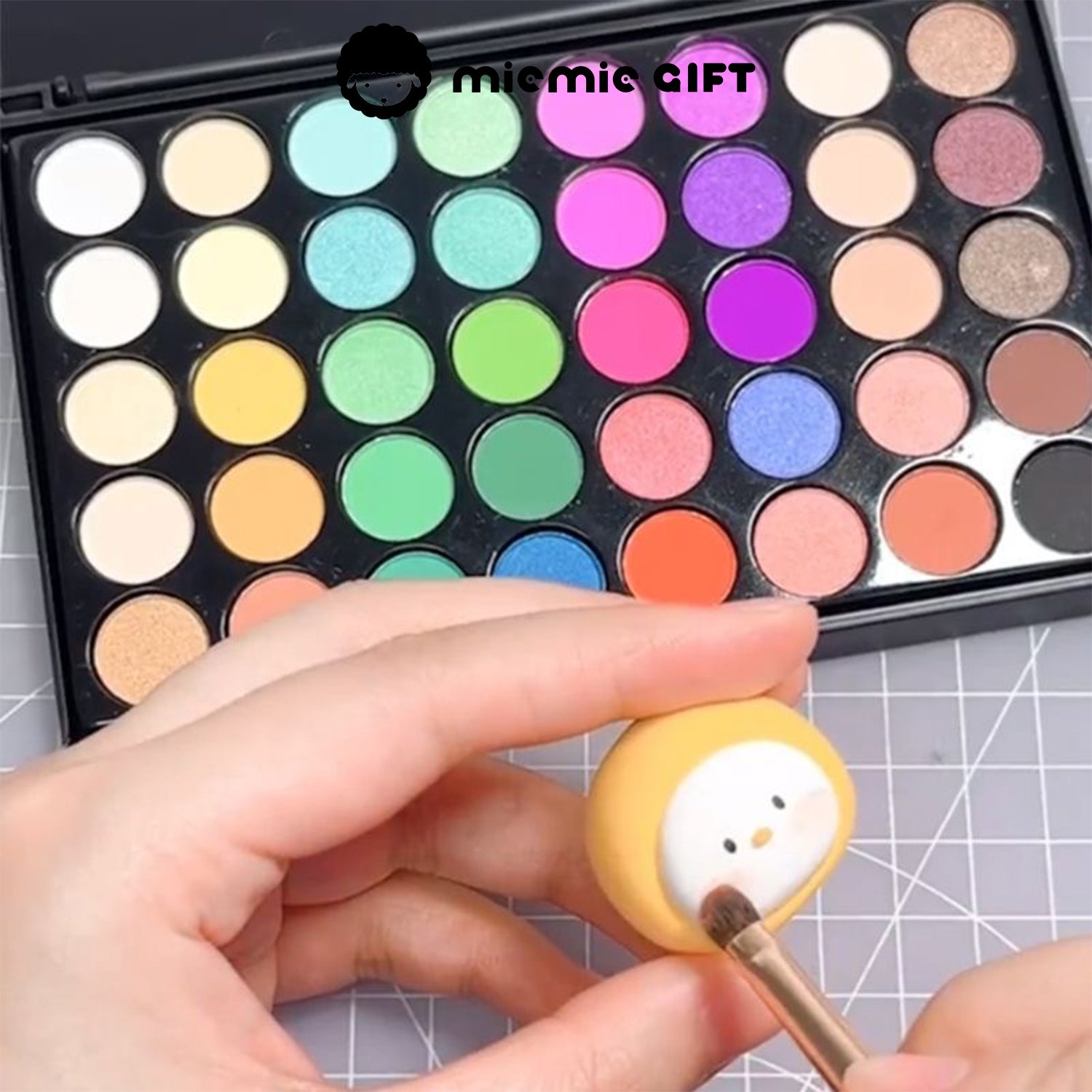 A hand using the MieMie Gift 40-Color DIY Chalk Powder Palette to add shading to a small clay figurine. The background features the Vivid Dream Palette with bright, bold colors, ideal for various DIY crafts.