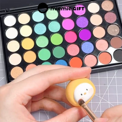 A hand using the MieMie Gift 40-Color DIY Chalk Powder Palette to add shading to a small clay figurine. The background features the Vivid Dream Palette with bright, bold colors, ideal for various DIY crafts.