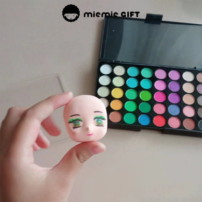 A hand holding a small doll head model while using the MieMie Gift 40-Color Vivid Dream Palette to add delicate facial coloring, particularly focusing on eyeshadow. This demonstrates the fine and detailed application of the chalk powder for doll customization.