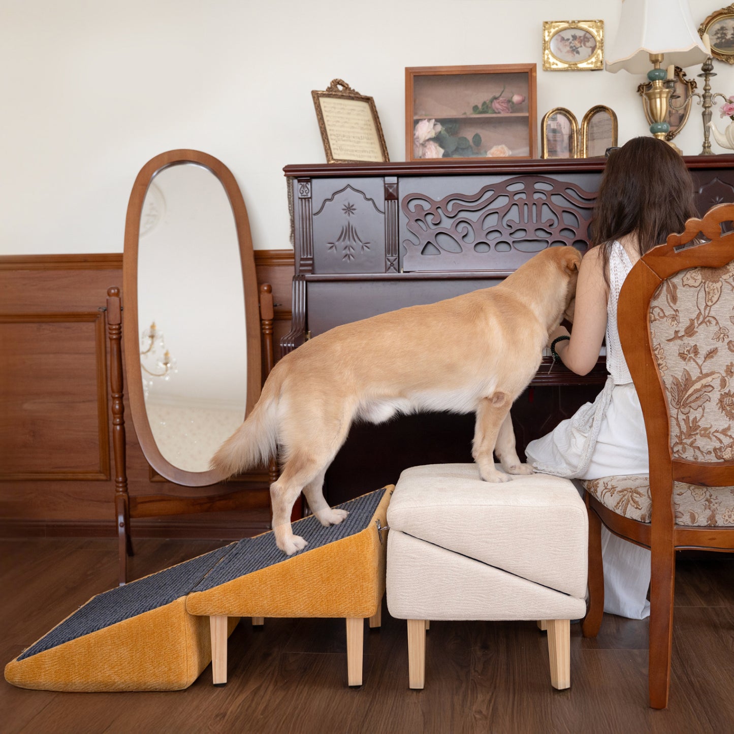 A golden retriever using a mustard-yellow dog ramp with a cream-colored ottoman to reach a woman sitting at a vintage dressing table. The dog stands beside the owner, creating a heartwarming moment of connection. The room features elegant wooden furniture, a tall mirror, and classical framed art, enhancing the cozy, timeless atmosphere. The ramp setup helps the pet safely climb up to interact with the seated person.