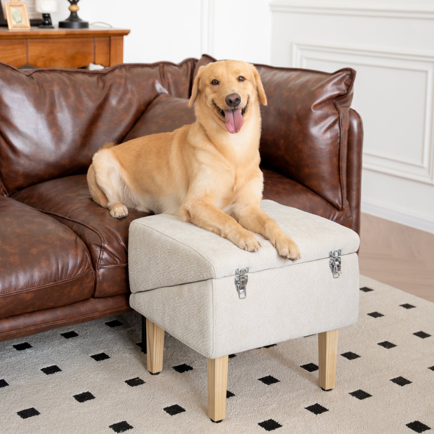Happy golden dog relaxing on the MieMie's Corner 3-in-1 dog ramp and ottoman seat in cream color, placed next to a brown leather sofa in a cozy living room. Multifunctional pet furniture used as a footstool and resting spot, featuring secure metal clasps and wooden legs.